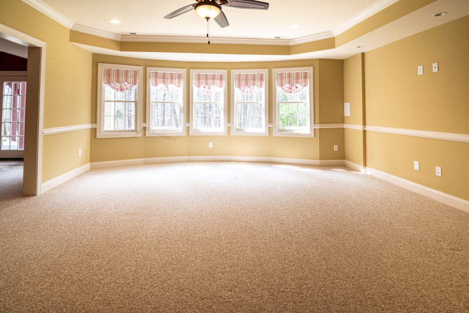 A spacious, well-lit living room featuring light-colored wooden flooring and a large, white L-shaped sofa positioned against the far wall, beneath a set of sheer white curtains covering a broad window