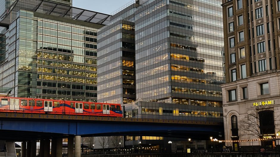 Urban scene in Limehouse featuring modern glass office buildings with reflective surfaces, a red and blue train traveling on an elevated railway track, and historic-style buildings with arched windows